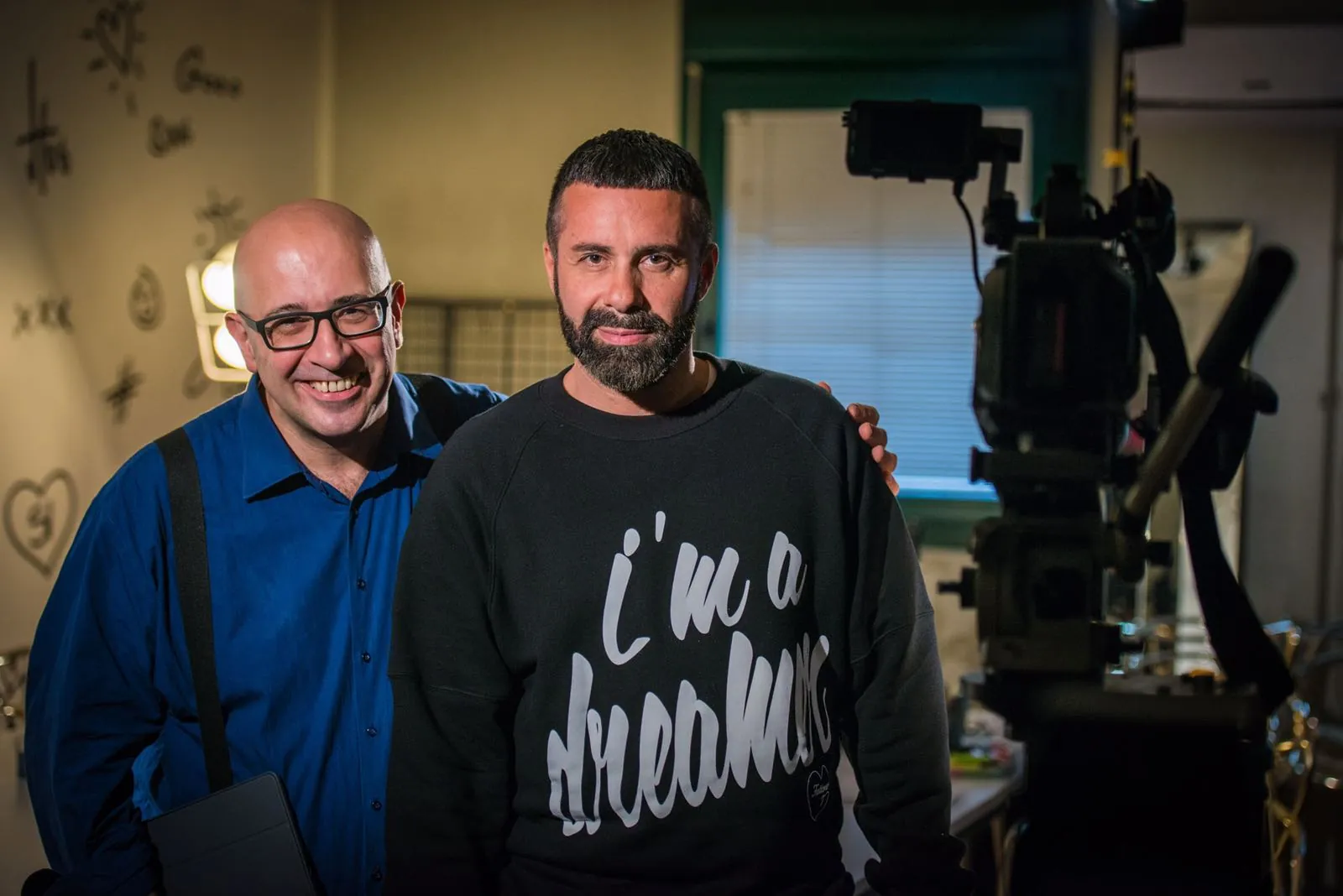Paolo Monesi sorride accanto a un artista in felpa su un set con macchina da presa