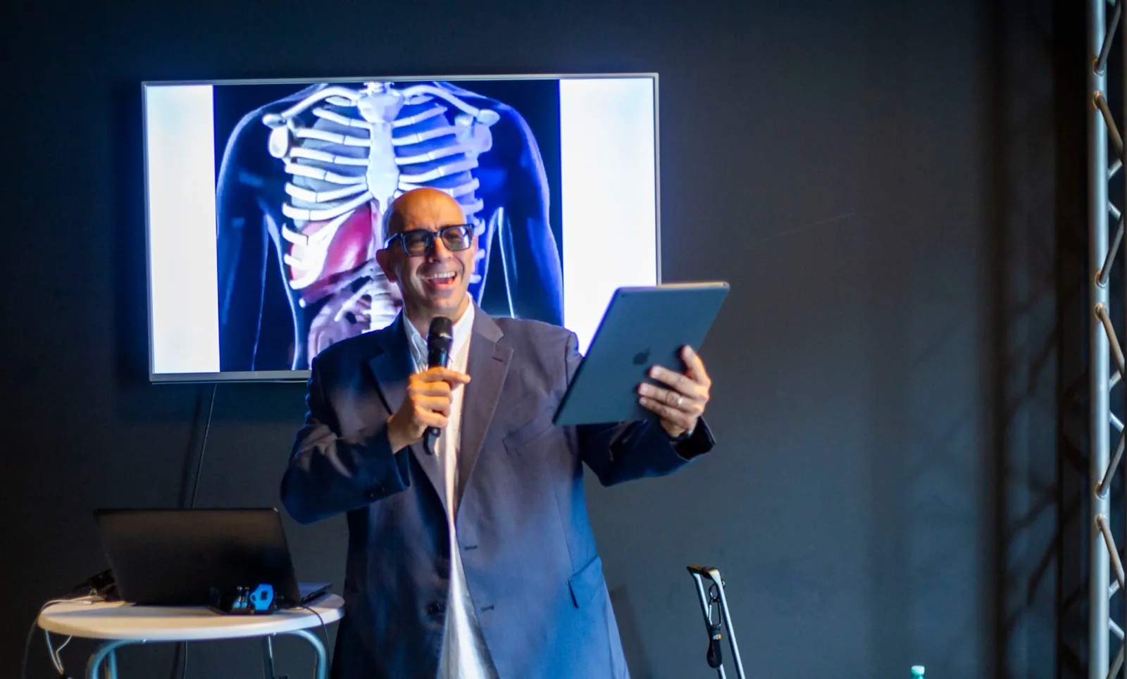 Paolo Monesi ride al microfono durante un intervento, con un tablet e uno schermo alle spalle